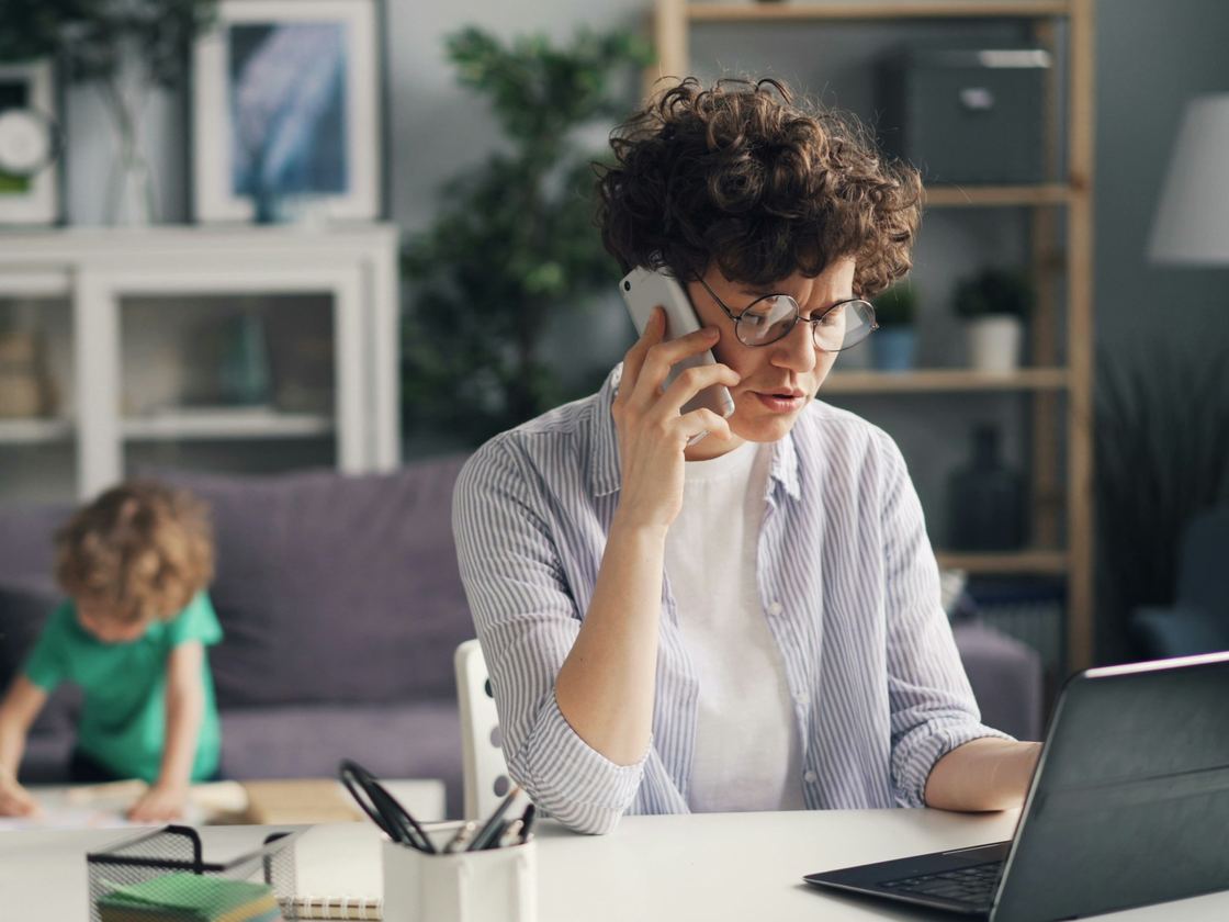 A woman on a phone call types on a laptop at a white desk while a young boy draws on the sofa behind her