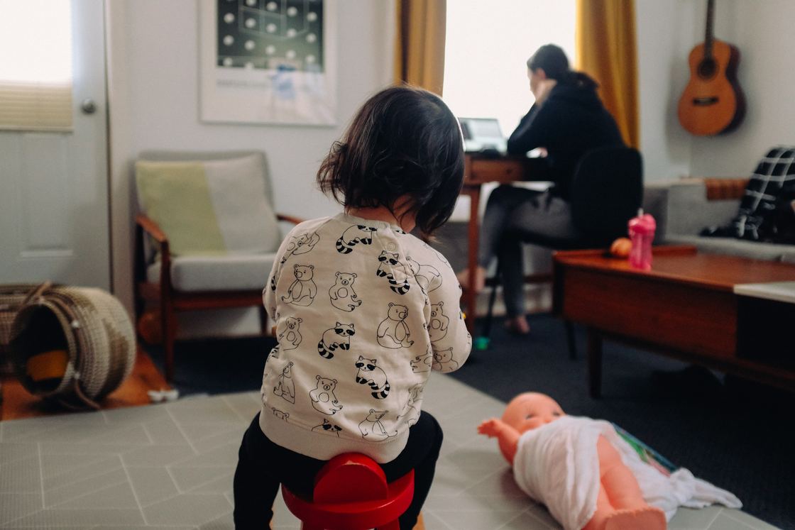 A toddler in a bear-print sweatshirt sits on a red stool while a woman works at a laptop by the window
