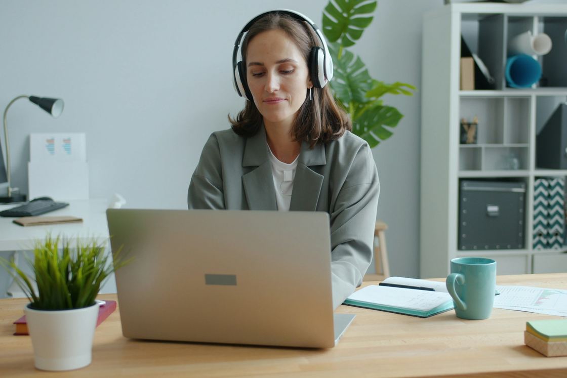A woman in a gray blazer and white headphones types on a laptop at a wooden desk