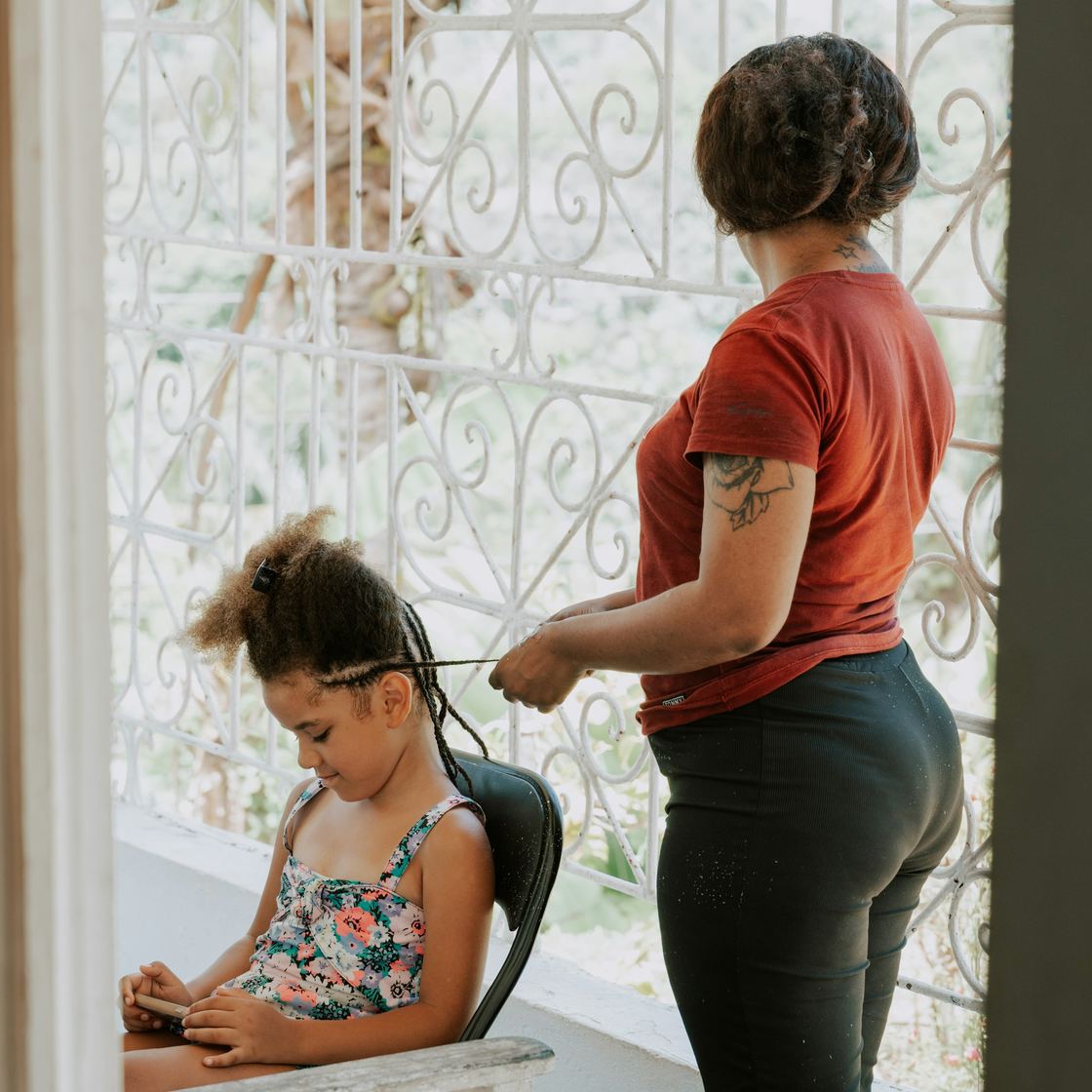 A woman braids a girl’s hair on a sunny balcony while the girl looks at a phone