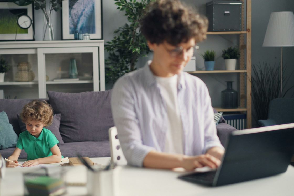 A woman types on a laptop at home while a curly-haired boy draws at a low table behind her